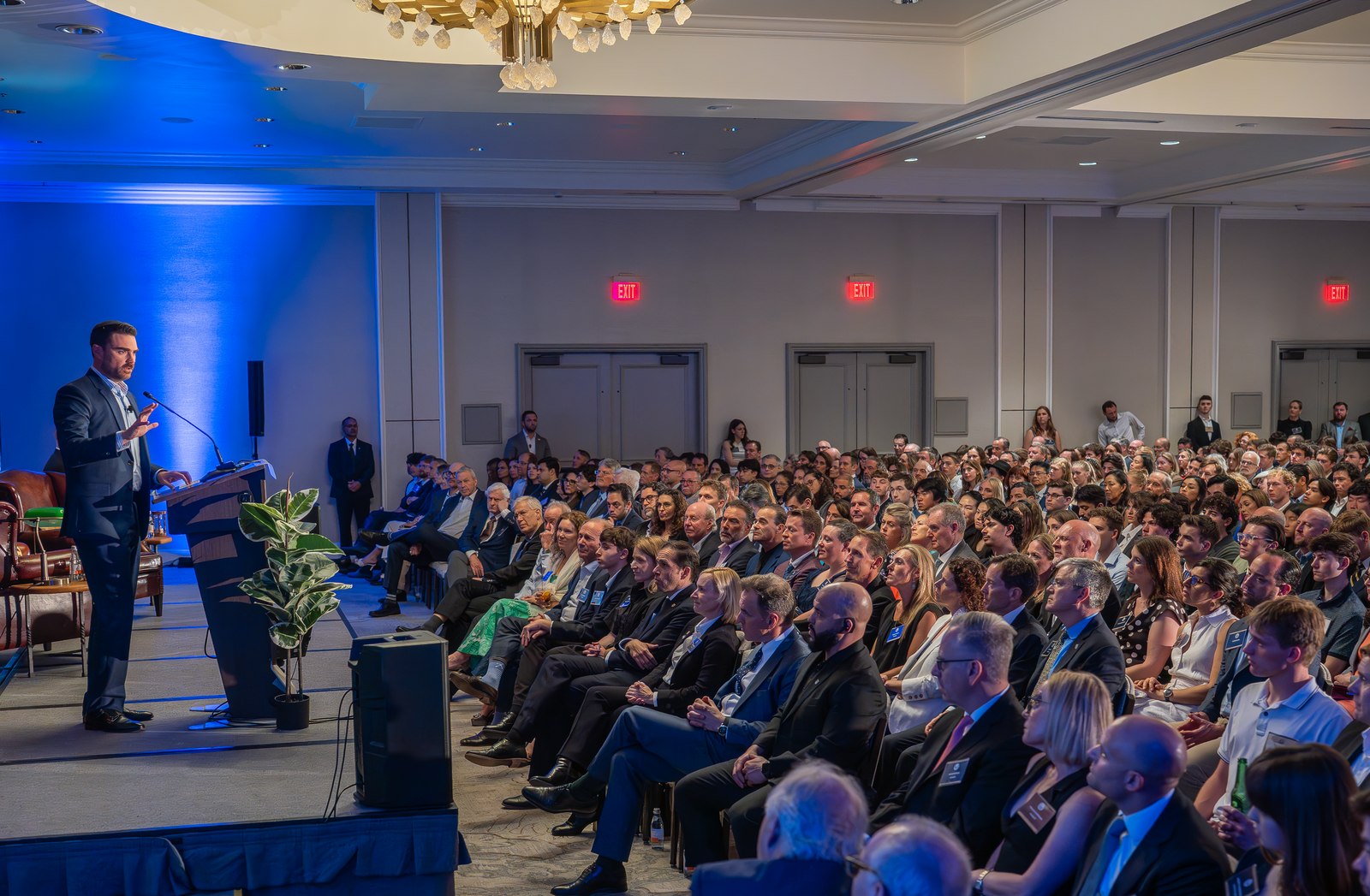 A speaker addressing a seated audience in a blue-lit ballroom.